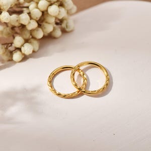 May include: Two gold hoop earrings with textured detailing. One earring has a braided design, the other a repeating pattern. The earrings are displayed on a light surface, suggesting jewellery or accessories.