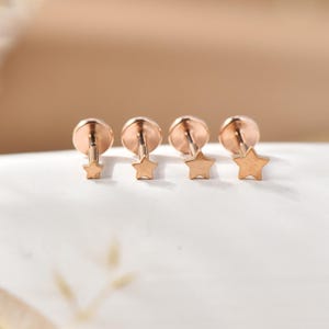 Könnte beinhalten: Vier roségoldfarbene Ohrringe in Sternform. Jeder Ohrring hat einen kleinen Sternanhänger, der an einem runden Stecker hängt. Die Ohrringe werden auf einer weißen Oberfläche mit unscharfem Hintergrund präsentiert.