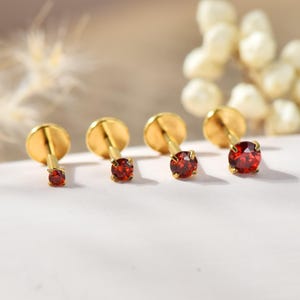 May include: Four gold-coloured stud earrings with red gemstones. The earrings are arranged in a row, with the gemstones increasing in size. The earrings have a flat, round backing and a post for piercing.