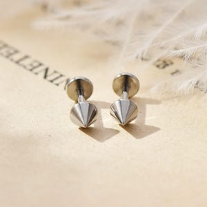 May include: A pair of silver-colored stud earrings. Each earring features a conical, pointed design. The earrings are displayed on a light-colored, textured surface, with a soft, blurred background.