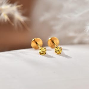 May include: A pair of gold-coloured stud earrings, each featuring a small, round, yellow gemstone. The earrings are set against a white surface with a soft, blurred background. The studs have a simple, elegant design.