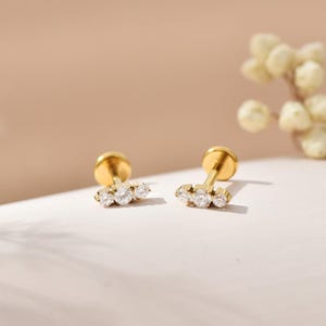 Könnte beinhalten: Zwei goldfarbene Ohrstecker, jeweils mit drei klaren, rund geschliffenen Edelsteinen. Die Ohrringe werden auf einer weißen Oberfläche mit einem weichen, neutralen Hintergrund präsentiert. Die Ohrstecker haben einen Stecker und eine flache Rückseite.