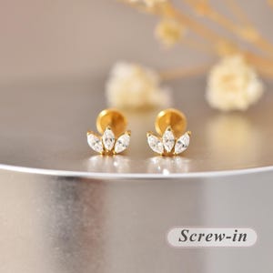 Peut inclure: Une paire de boucles d'oreilles à tige dorées avec trois pierres claires taille marquise disposées en éventail. Les boucles d'oreilles ont un mécanisme de fermeture à vis. Le texte "Screw-in" est visible dans le coin inférieur droit.