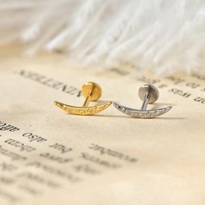 Könnte beinhalten: Zwei halbmondförmige Ohrringe, einer goldfarben und einer silberfarben, werden auf einem Vintage-Papierhintergrund gezeigt. Die Ohrringe haben eine strukturierte Oberfläche und einen Steckerverschluss. Der goldene Ohrring ist links, der silberne rechts.