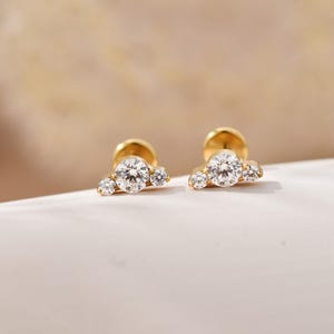 Könnte beinhalten: Ein Paar goldene Ohrstecker, jeweils mit drei klaren, runden Edelsteinen. Der größte Stein befindet sich in der Mitte, flankiert von zwei kleineren Steinen. Die Ohrringe haben eine polierte Oberfläche, vor einem weichen, neutralen Hintergrund.