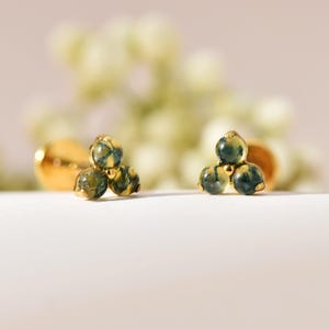 May include: A pair of stud earrings featuring three small, round, moss-green patterned stones arranged in a triangular cluster, set in a polished gold-tone metal base with a secure post backing for attachment to the earlobe.