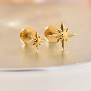 May include: A pair of gold-colored star-shaped stud earrings. Each earring features a detailed eight-pointed star design. The earrings are set on a post with a round backing, and are displayed on a light-colored surface.