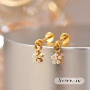 Könnte beinhalten: Ein Paar goldfarbene Schraub-Ohrstecker mit einem hängenden Blumenanhänger. Jede Blume besteht aus sechs kleinen, klaren, rund geschliffenen Steinen, die kreisförmig angeordnet sind. Die Ohrringe haben ein poliertes Gold-Finish und einen Schraubverschluss.