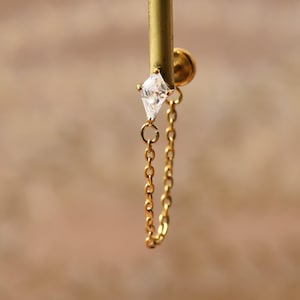 Könnte beinhalten: Ein goldfarbenes Körperschmuckstück mit einem rautenförmigen, klaren Edelstein in einer Metallfassung, verbunden mit einer feinen goldfarbenen Kette, die zum Stecker zurückführt. Der Schmuck hat einen flachen, runden Verschluss und ein poliertes Finish.