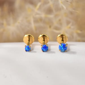 Könnte beinhalten: Drei goldfarbene Ohrstecker mit runden, blauen Opalsteinen. Die Ohrringe sind auf einer weißen Oberfläche mit unscharfem Hintergrund dargestellt. Die Opalsteine haben eine lebendige, schillernde Qualität.