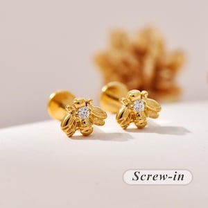 May include: A pair of gold bee-shaped stud earrings featuring a clear round gemstone set in the center of each bee body. The earrings have a screw-in backing mechanism. The text "Screw-in" is visible in the lower right corner of the image.