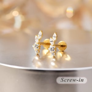 Könnte beinhalten: Ein goldfarbener Schraub-Ohrstecker mit einer Anordnung aus klaren, facettierten Steinen in einem vertikalen, blattartigen Muster mit einem sternförmigen Zentrum. Das Design umfasst Steine im Marquise- und Rundschliff auf einer Metallbasis.