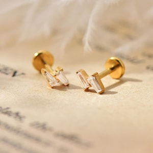 Könnte beinhalten: Zwei goldfarbene Ohrstecker mit geometrischem Design. Jeder Ohrring hat eine kleine, runde Rückseite und einen Stecker. Die Vorderseite jedes Ohrrings hat ein Zickzackmuster aus klaren, rechteckigen Steinen.