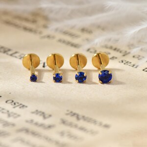 May include: Four gold-colored stud earrings with round, bright blue gemstones. The earrings are arranged in a row, with the gemstones increasing in size from left to right. The earrings are displayed on a vintage paper background.