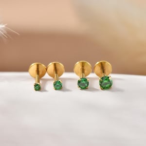 Könnte beinhalten: Vier goldfarbene Ohrringe mit flacher Rückseite und unterschiedlich großen, runden, smaragdgrünen Edelsteinen. Die Ohrringe werden auf einer weißen Oberfläche mit einem weichen, verschwommenen Hintergrund präsentiert. Die Ohrringe sind für Ohrpiercings konzipiert.