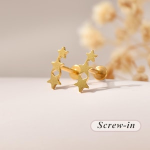 May include: A pair of gold-toned stud earrings featuring a vertical arrangement of three connected star shapes with a small diamond-shaped cutout in the centre. The earrings have a screw-in back mechanism as indicated by the text Screw-in.
