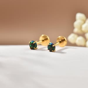 May include: Two gold-colored stud earrings with iridescent, dark green and blue gemstones. The earrings are displayed on a white surface with a soft, neutral background. The gemstones are round and set in a simple, elegant design.