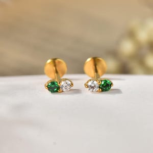 Könnte beinhalten: Ein Paar goldfarbene Ohrstecker, die jeweils mit zwei kleinen, nebeneinander liegenden, rund geschliffenen Steinen besetzt sind. Ein Stein ist leuchtend smaragdgrün, der andere ist ein klarer, farbloser Stein. Die Ohrstecker haben einen flachen Verschluss.