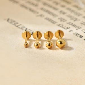 Könnte beinhalten: Vier goldfarbene Ohrstecker mit kugelrunden Enden, präsentiert auf einer hellen Oberfläche. Die Ohrringe sind in verschiedenen Größen erhältlich, mit kleinen, mittleren und großen Kugeldesigns. Die Ohrringe haben eine flache Scheibenrückseite.