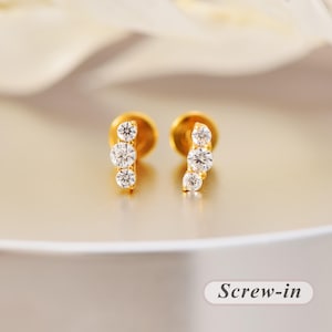 Könnte beinhalten: Ein Paar goldene Ohrstecker, jeweils mit drei runden, klaren Steinen in einem vertikalen Bogen angeordnet. Die Ohrringe verfügen über einen Schraubverschluss. Der Text "Screw-in" ist in der unteren rechten Ecke des Bildes zu sehen.