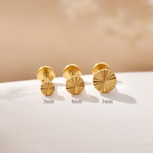 Könnte beinhalten: Drei goldfarbene Ohrstecker mit strukturierten, sonnenstrahlenartigen Oberflächen. Die Ohrringe haben einen Durchmesser von 3 mm, 4 mm und 5 mm. Die Stifte und Rückseiten sind ebenfalls goldfarben. Auf weißer Oberfläche präsentiert.