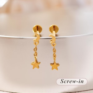 Könnte beinhalten: Ein Paar goldfarbene Schraub-Ohrstecker mit einem Mondsichel-Anhänger, der an einer kurzen Kette mit einem kleinen Stern-Anhänger befestigt ist. Die Ohrringe haben einen flachen, runden Verschluss und eine polierte metallische Oberfläche.