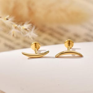 May include: A pair of gold-toned stud earrings featuring a smooth, curved crescent moon shape. Each earring has a flat circular backing attached to a straight post, designed for piercing. The metal surface is polished and reflects light evenly.