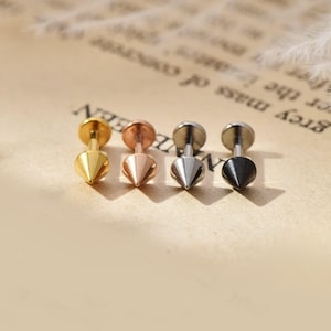 May include: Four stud earrings with cone-shaped spikes. The earrings are gold, rose gold, silver, and black. The earrings are displayed on a light brown surface.