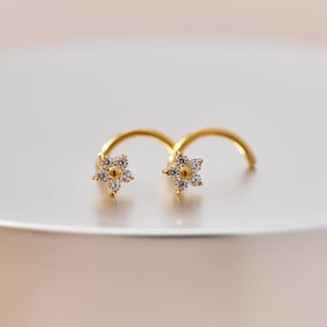 Könnte beinhalten: Zwei goldfarbene Nasenstecker, jeweils mit einem kleinen, blütenförmigen Design. Die Blüte hat eine zentrale goldfarbene Perle, umgeben von klaren, funkelnden Kristallen. Die Stecker sind für einen bequemen Sitz gebogen.