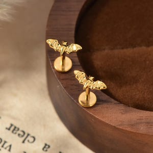May include: A pair of gold-toned stud earrings featuring a bat-shaped design with outstretched wings. Each earring has a flat circular backing and a post, crafted with a polished metallic finish that reflects light across the detailed bat silhouette.