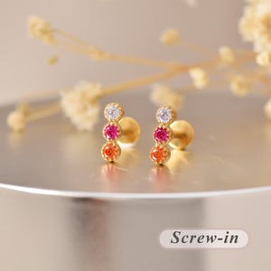 May include: A pair of gold stud earrings featuring a vertical row of three round-cut stones in a gradient of white, pink, and orange. Each earring has a screw-in back mechanism. The design displays a polished gold setting holding the stones.