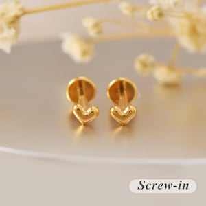 Peut inclure: Une paire de boucles d'oreilles à tige dorées avec un motif en forme de cœur et un fermoir plat circulaire. Le modèle est identifié comme étant à visser, avec le texte "Screw-in" clairement visible dans le coin inférieur droit de l'image.