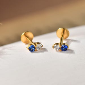 Peut inclure: Une paire de boucles d'oreilles à dos plat de couleur dorée, chacune ornée de deux petites pierres rondes côte à côte. Une pierre est d'un bleu vif, tandis que l'autre est transparente et incolore. Elles présentent un design à tige.