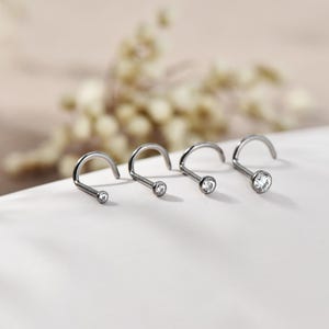 Könnte beinhalten: Vier silberne Nasenstecker mit gebogenen Stiften und klaren, runden Edelsteinen. Die Stecker sind in einer Reihe angeordnet, jeder mit einem etwas größeren Edelstein als der vorherige. Der Hintergrund ist in einer weichen, neutralen Farbe gehalten.