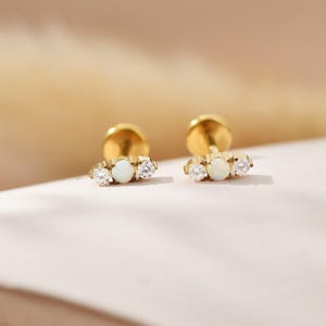 May include: A pair of gold stud earrings, each with an opal gemstone centre and two clear cubic zirconia stones. The earrings are set on gold posts and backs. The background is a soft, neutral colour.
