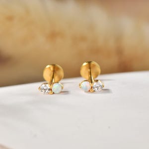 Könnte beinhalten: Ein Paar goldene Ohrstecker, die jeweils mit zwei kleinen, nebeneinander liegenden runden Steinen besetzt sind. Ein Stein ist ein klarer, facettierter weißer Edelstein, der andere ein glatter, schillernder Opal. Die Ohrstecker haben einen flachen Verschluss.
