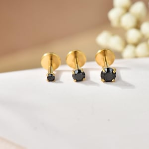 Könnte beinhalten: Drei goldfarbene Ohrstecker mit schwarzen Edelsteinen. Die Ohrringe sind in einer Reihe angeordnet und zeigen die unterschiedlichen Größen der schwarzen Steine. Die Ohrstecker haben ein schlichtes, elegantes Design mit einem runden Edelstein in einer goldenen Lünette.