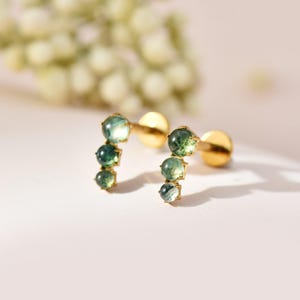 May include: A pair of gold-tone stud earrings, each featuring three small, round, translucent green moss agate stones arranged in a curved line. The earrings have a flat circular backing and a post mechanism for secure attachment to the earlobe.