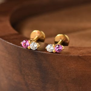 May include: A pair of gold stud earrings, each featuring two round-cut stones set side-by-side. One stone is a vibrant pink, while the other is a clear, colorless stone. The earrings have a flat, circular backing post design for secure placement.