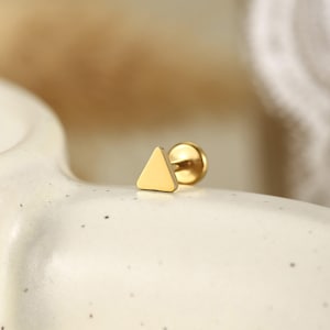 Könnte beinhalten: Ein goldfarbener Ohrstecker mit einem flachen, glatten, dreieckigen Vorderteil und einem runden Verschluss. Die Metalloberfläche hat ein poliertes, reflektierendes Finish und zeigt ein schlichtes geometrisches Design mit abgerundeten Kanten.