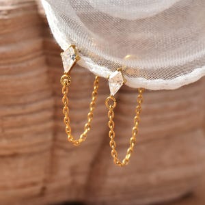 May include: A pair of gold chain earrings. Each earring has a diamond-shaped, clear gemstone at the top, linked to a delicate chain that hangs down. The earrings are shown against a soft, white fabric background.