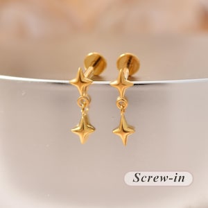 Könnte beinhalten: Ein Paar goldfarbene Schraubohrringe mit einem vierzackigen Sternmotiv. Jeder Ohrring besteht aus einem festen Stern oben und einem hängenden Sternanhänger, der an einem kleinen Ring befestigt ist. Der Text Screw-in ist in der Ecke sichtbar.