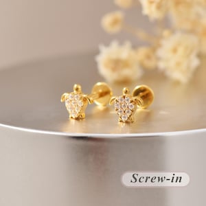 May include: A pair of gold-toned stud earrings shaped like sea turtles, each featuring a shell encrusted with small, clear, sparkling stones. The earrings have a flat back post design and include the text Screw-in on the lower right corner.