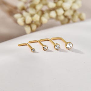 May include: Four gold nose studs, each with a clear, round gemstone. The studs are arranged in a row, with varying gemstone sizes. The L-shaped posts are visible. The background is a soft, neutral colour.