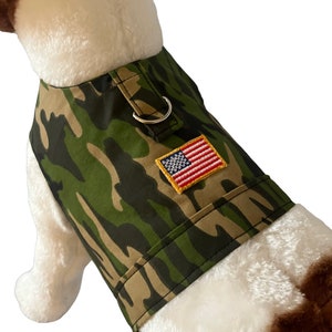 May include: Camouflage dog harness with a small American flag patch. The harness is green and brown with a silver D-ring.