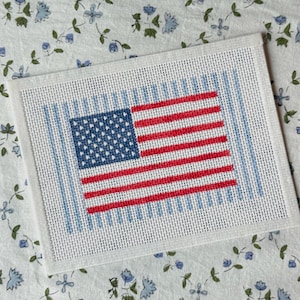 May include: A rectangular needlepoint canvas featuring a stitched American flag design. The flag consists of a blue canton with white stars and alternating red and white horizontal stripes, centered against a background of vertical light blue lines.