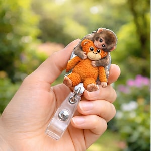 May include: A badge reel featuring a brown monkey hugging an orange teddy bear. The badge reel has a clear plastic clip and a metal retractable cord. The background is a blurred view of green foliage.