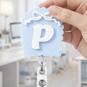 May include: A light blue badge reel with a white scalloped edge and a white bow design at the top. The letter "P" is in white. The reel has a clear plastic clip and a metal clasp.