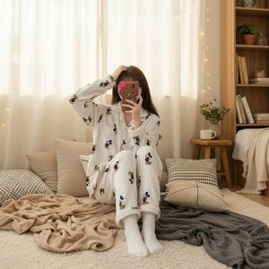 May include: White pajama set with a button-down top and pants, featuring a repeating Mickey Mouse pattern in black, red, and yellow. The pajamas are made of a soft, plush material. The person is holding a brown phone case with a pink bow.