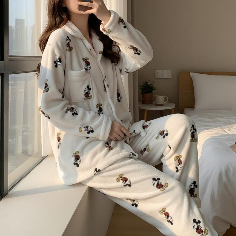 May include: White fleece pajama set featuring a repeating Mickey Mouse pattern in black, red, yellow, and tan. The long-sleeved top has a button-down front and a chest pocket. The pants are loose-fitting.